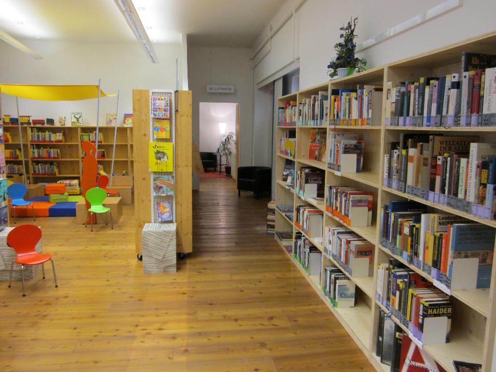 Bibliothek innen rechts: Kinderbereich am linken Teil des Fotos, dann ein Regal und ein Gang und rechts am Foto ein Regal an der Wand